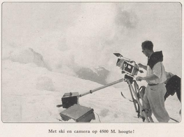 Cameraman met een grote camera op een statief op een besneeuwde berghelling, onderschrift: Met ski en camera op 4500 M. hoogte!